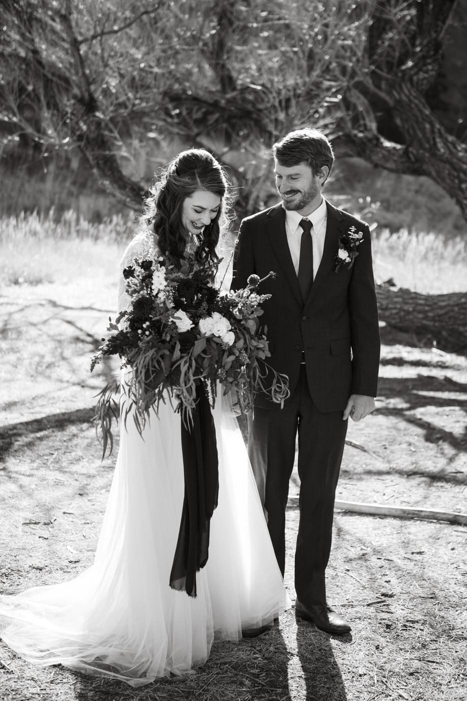 bride and groom posed outdoors in the grass as he looks at her and she looks at her bouquet