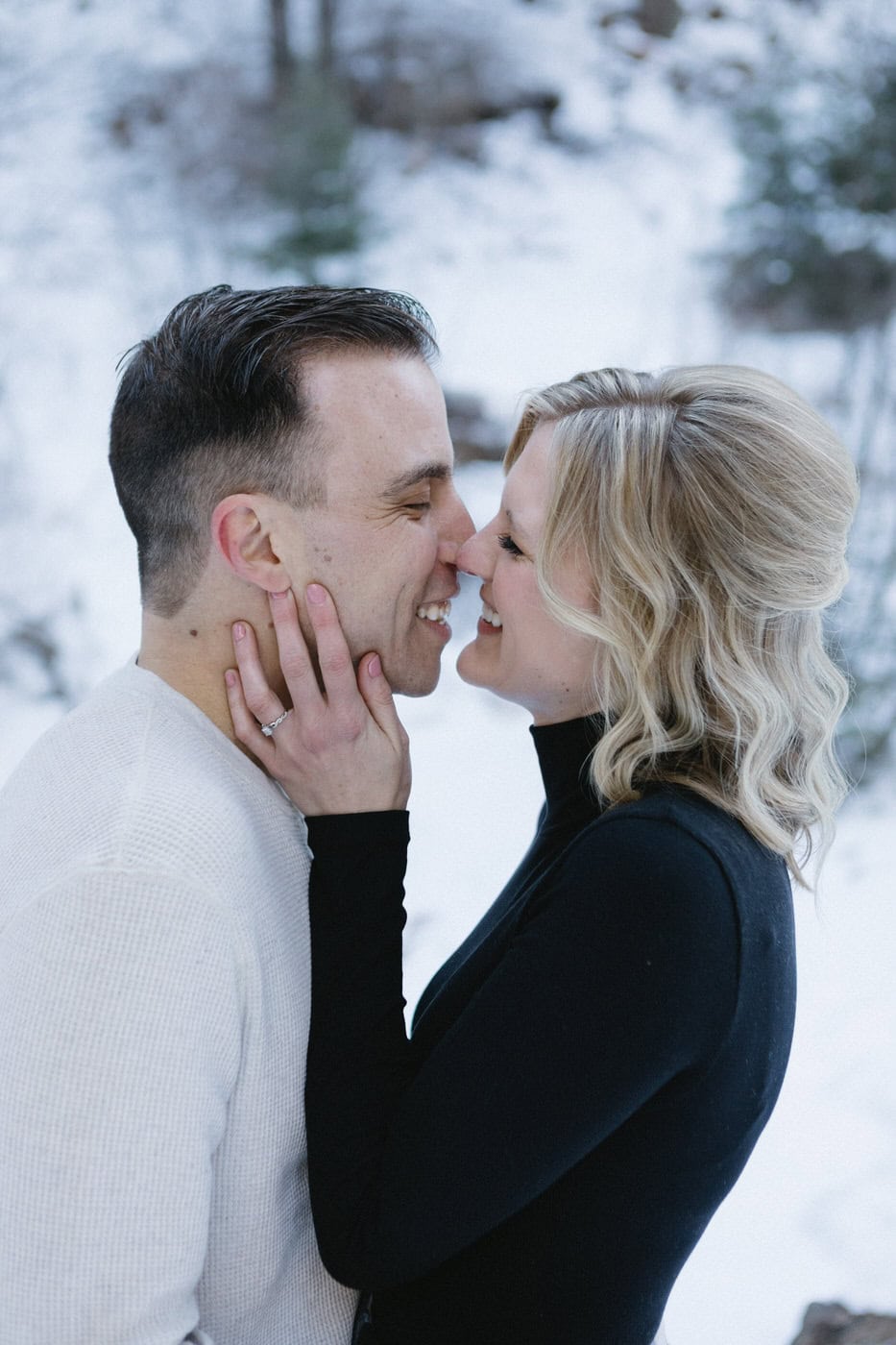 An engaged couple stand nose to nose and she has his hand on his cheek.